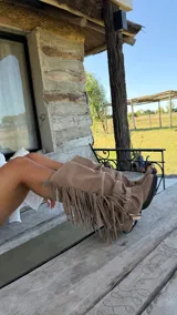 Botas de caña media confeccionadas en gamuza, con diseño holgado y detalle de flecos laterales con cadena metálica. Cuentan con suela plana y estilo western.