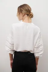 Camisa de color crudo con diseño romántico, cuello redondo con cordón ajustable terminado en borlas, y mangas tres cuartos abullonadas. Presenta detalles de bordados calados verticales en el frente y horizontales en las mangas.