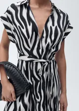 Vestido camisero midi de tejido fluido con estampado de cebra. Presenta cuello camisero, cierre frontal de botones, mangas cortas y lazo ajustable en la cintura para entallar la silueta.