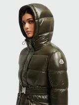 Campera larga tipo puffer de Moncler, color verde oliva con acabado brillante. Presenta capucha integrada, cierre frontal con cremallera y cinto ajustable en la cintura con hebilla metálica.