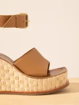 Sandalia de plataforma color marrón claro, realizada en cuero vacuno con suela de madera forrada en rafia. El interior es en cuero vacuno. Tiene hebilla en tobillo para regular su calce.