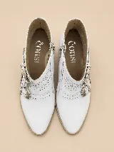 Botas cortas de cuero blanco con taco cuadrado bajo y punta fina. Cuentan con cierre lateral y tres tiras cruzadas con hebillas metálicas y tachas decorativas.