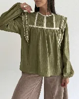 Blusa de algodón color verde oliva, con cuello redondo, mangas largas abullonadas con puños elásticos y detalles de puntilla y bordados florales en el frente y hombros.