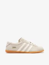Championes Adidas Gazelle Lo Pro W de perfil bajo, confeccionados en gamuza color gris claro, con las tres tiras características en tono crudo y suela de goma translúcida. Incluye el nombre del modelo y la marca en letras doradas en el lateral.
