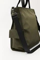 Bolso tipo tote color negro, con doble asa superior y correa desmontable. Presenta un bolsillo frontal con solapa, bolsillos laterales y un detalle colgante decorativo con tiras de colores.