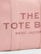 Bolso tipo tote de lona color negro con letras blancas bordadas en el frente que dicen 'THE TOTE BAG MARC JACOBS'. Cuenta con asas superiores cortas y una correa de hombro ajustable y extraíble.