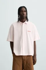 Camisa de algodón color rosa pálido, corte boxy fit, con cuello solapa, manga corta y abotonadura frontal. Presenta un texto bordado en contraste de color rojo en el delantero.