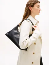 Cartera tipo hobo Tommy Hilfiger de acabado liso en color negro, con cierre de cremallera superior, asa corta de mano y correa tejida ajustable y extraíble en blanco y negro. Incluye detalle de logo metálico en relieve en la parte delantera y herrajes dorados.