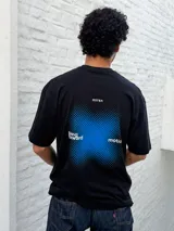 Remera negra de corte oversize con cuello redondo y manga corta. Presenta un pequeño estampado blanco con texto y un cuadrado difuminado azul en el pecho.