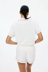 Pijama de dos piezas confeccionado en mezcla de viscosa, compuesto por una camisa de manga corta con cuello abierto, botones frontales y bolsillo superior, y un short con elástico en la cintura. Ambos presentan ribetes decorativos en contraste.