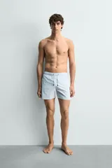 Bañador de hombre de largo medio, color celeste claro con rayas blancas horizontales. Presenta cintura elástica ajustable con cordón, bolsillos frontales y bolsillos traseros de plastrón. Incluye forro interior.