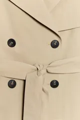 Trench corto de color beige, con cuello de solapa, manga larga con hombreras y trabillas con botones. Cuenta con cierre frontal cruzado con botones oscuros y cinturón ajustable del mismo tejido.