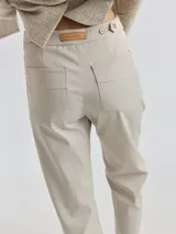 Pantalon de corte cigarette color beige, con cierre frontal, bolsillos laterales y detalle de botón decorativo en la cintura. Presenta costuras reforzadas con hilo rojo en los extremos de los bolsillos.
