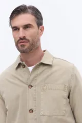 Sobrecamisa de canvas color beige con efecto lavado, de estructura firme y manga larga. Presenta cuello clásico, cierre frontal con botones y un bolsillo frontal en el pecho.