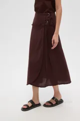 Falda larga de algodón color marrón oscuro, de corte envolvente (wrap skirt) con abertura lateral y borde deshilachado. Se ajusta con un lazo lateral que pasa por argollas metálicas.