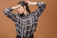 Camisa de manga larga con estampado a cuadros, cuello alto y cierre frontal con botones. Presenta un diseño fluido con lazo decorativo en el cuello y puños abotonados.