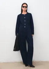 Mono largo de denim color azul oscuro, con cuello redondo, manga larga y cierre frontal de botones metálicos. Presenta corte recto y amplio en las piernas.