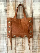 Cartera tipo tote de cuero color marrón, con diseño estilo boho. Presenta detalles de tachas metálicas circulares grandes y flecos laterales largos. Cuenta con doble asa superior para llevar al hombro.