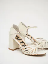 Sandalias de tacón cuadrado de 8 cm, confeccionadas en charol color crema. Presentan un diseño de tiras finas entrelazadas en la parte frontal y una correa ajustable con hebilla metálica en el tobillo.