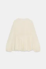 Blusa de viscosa color rosado con cuello redondo, manga larga abullonada con puños elásticos y cierre frontal con lazadas. Presenta un diseño con volantes en el bajo y corte asimétrico.