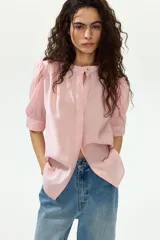 Blusa color rosado con cuello mao y botones ocultos en la parte delantera. Tiene canesú de doble capa con frunces adelante y atrás, mangas cortas abullonadas y puños con abertura y botón.