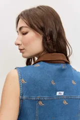 Chaleco de denim azul sin mangas, con cierre frontal de botones metálicos, bolsillos con solapa en el pecho y cuello camisero marrón de pana. Presenta bordados pequeños dispersos en color marrón.