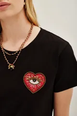 Remera negra de algodón con cuello redondo y manga corta. Presenta un aplique bordado en el pecho con forma de corazón rojo que contiene un ojo detallado con mostacillas y bordados.