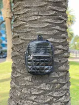Mochila de cuero sintético color negro con diseño combinado: parte superior con textura de reptil y parte inferior con diseño acolchado capitoneado. Cuenta con bolsillo frontal con cierre metálico y tirador decorativo.