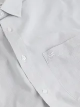 Camisa de manga larga blanca con finas rayas verticales grises. Presenta cuello clásico, cierre frontal de botones y un bolsillo de parche en el pecho con logo bordado.