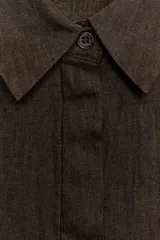 Camisa de lino color marrón oscuro con cuello solapa, manga por debajo del codo y cierre frontal con botones ocultos por solapa. Presenta bolsillos de plastrón con solapa en el pecho y trabillas en los hombros.