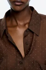 Camisa oversize de tejido fantasía marrón oscuro, con minipiedras distribuidas por toda la prenda. Presenta cuello camisero, botones frontales, bolsillos de plastrón en el pecho y mangas largas con puño abotonado.