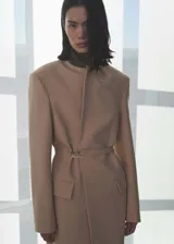 Abrigo largo de diseño oversize en color beige, confeccionado en tejido mezcla de lana. Presenta cuello con solapas, hombros estructurados y pronunciados, manga larga y dos bolsillos laterales con solapa. Incluye un cinturón fino ajustable en la cintura y abertura posterior en el bajo.