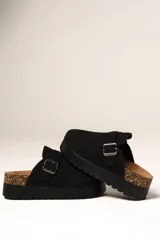 Zueco de plataforma color negro, con capellada de gamuza sintética y detalle de hebilla metálica ajustable en el empeine. Presenta una base de corcho en el talón y suela dentada de goma.