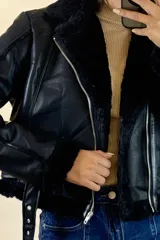 Campera tipo aviador de cuero sintético negro con interior y detalles de piel sintética al tono en cuello, puños y ruedo. Presenta cierre frontal cruzado y cinturón ajustable con hebilla metálica en la cintura.
