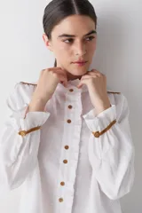 Camisa blanca de lino con cuello alto tipo gorguera, cierre frontal con botones de madera y detalles de ribetes en color mostaza en hombros y puños. Presenta volados en la botonera, cuello y puños.