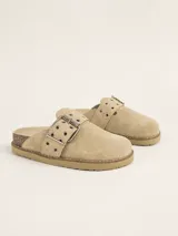 Zueco de gamuza color beige, con diseño de punta cerrada y talón descubierto. Presenta una tira ajustable sobre el empeine con hebilla metálica y ojales decorativos. Cuenta con una suela de corcho y base de goma antideslizante.