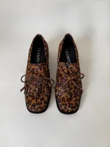 Mocasín de cuero marrón con textura granulada, diseño de horma cuadrada, detalle de flecos y lazo decorativo en el empeine, montado sobre una suela y taco bajo de color negro.