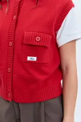 Chaleco de punto rojo con cierre frontal de botones, cuello redondo y dos bolsillos frontales con solapa. Presenta un diseño de tejido acanalado en los bordes y un pequeño parche decorativo en el pecho.