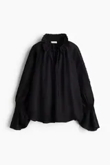 Blusa oversized color negro en tejido liviano de mezcla de lino y viscosa, con cuello elevado plisado y abertura en V con tiras finas. Mangas raglán extralargas con detalles bordados tono sobre tono y puños acampanados con pliegues.