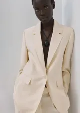 Blazer de diseño recto y estilo sastrero, color beige, confeccionado en mezcla de lyocell y poliéster. Cuenta con cuello en pico con solapas de muesca, manga larga, bolsillos delanteros con solapa y cierre frontal de un botón.
