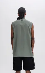 Camisa sin mangas color verde militar, con cuello, cierre frontal de botones y un bolsillo de parche con tapa en el pecho izquierdo. Presenta un calce oversize.