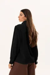 Camisa negra de textura sutil, con cuello alto tipo mao, cierre frontal con botones y detalle de volado lateral. Presenta mangas largas con puños con volados.