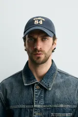 Gorra de seis paneles azul marino confeccionada en tejido de algodón, con bordado frontal en tono contrastante con la leyenda "button 84".