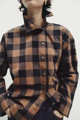 Camisa de manga larga con estampado de cuadros en tonos marrón y negro. Presenta un diseño cruzado con cuello vuelto y cierre frontal de botones, con un corte asimétrico en el bajo.
