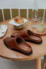 Mules de cuero color suela, con diseño abierto y tira frontal decorada con tachas metálicas circulares. Presentan punta cuadrada y suela plana.