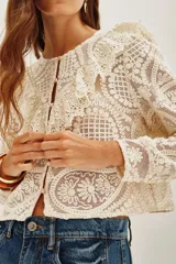 Saco corto de encaje color crema, con cuello redondo, mangas largas y volados en los hombros. Cierre frontal con botones dorados.