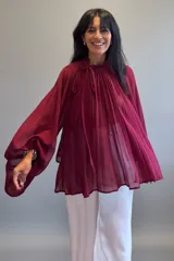 Blusa plisada de color bordó con diseño amplio y fluido. Presenta cuello alto con lazo para anudar, mangas largas con gran volumen y una textura plisada en toda la prenda.
