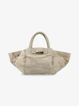 Cartera de diseño amplio y estructurado, confeccionada en gamuza color beige. Presenta asas cortas, laterales con volumen y un cierre frontal con detalle metálico dorado.