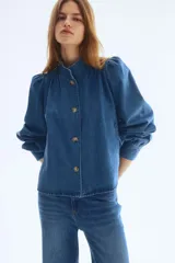 Campera de sarga de mezclilla azul, con cuello elevado, cierre frontal de botones y canesú fruncido. Tiene mangas largas abullonadas con puños ajustados y bolsillos laterales insertados.