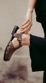 Sandalia tipo cangrejera de cuero color marrón, con diseño de tiras entrelazadas, hebilla lateral ajustable y suela plana de goma.
